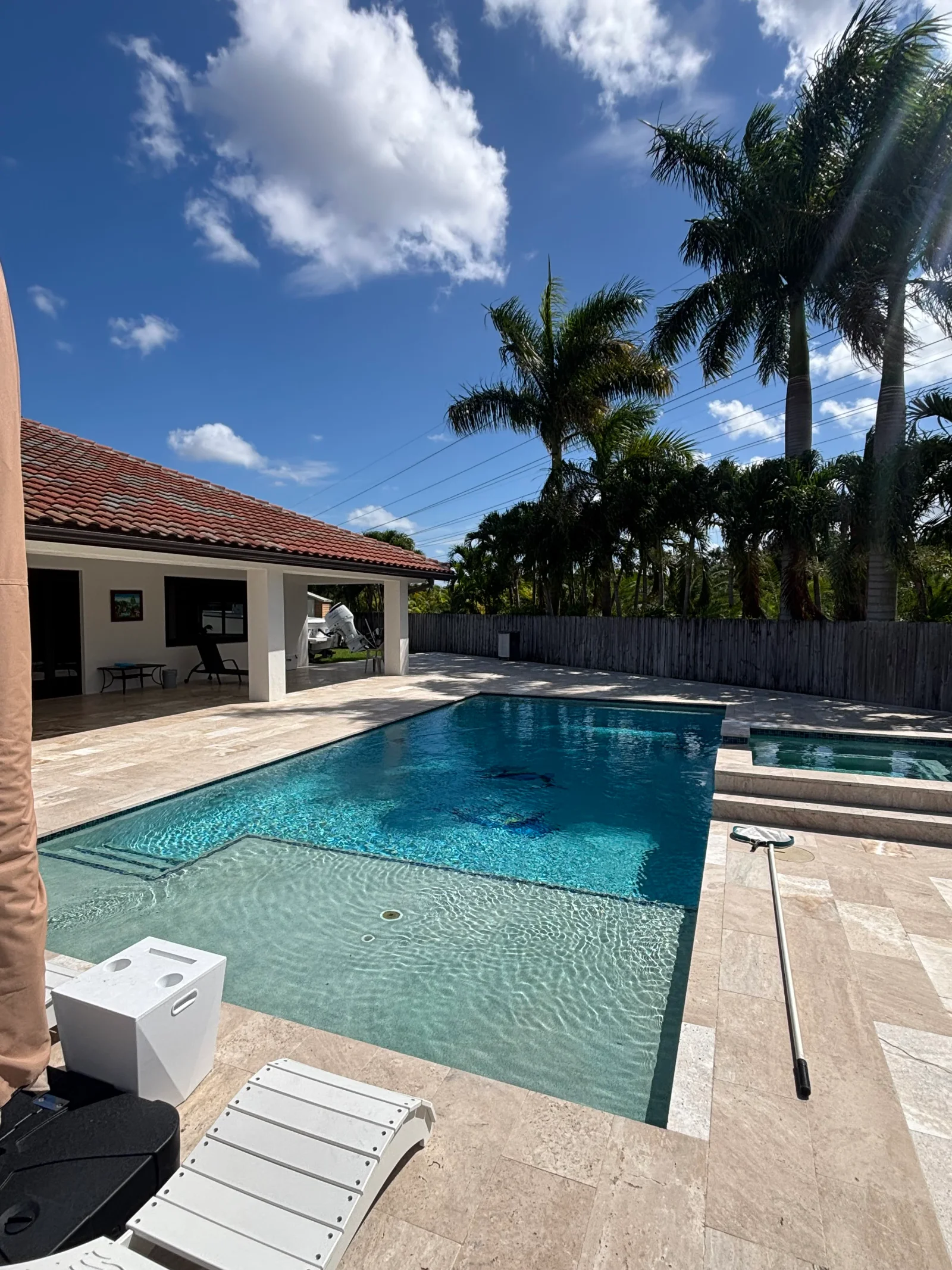 In-ground pool with travertine deck, covered patio, and wooden fence surrounding residential backyard